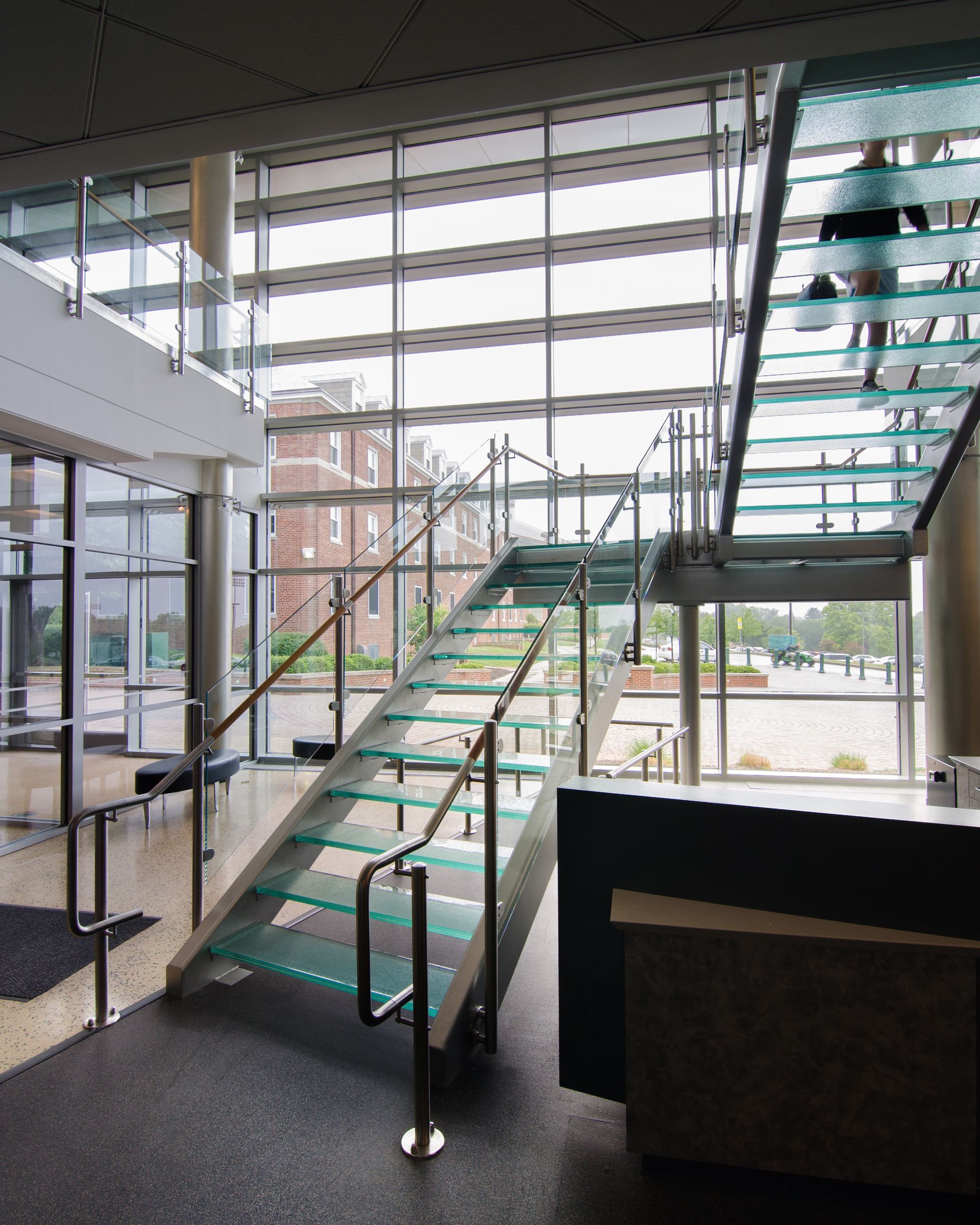 Modern lobby interior with a glass staircase, large windows, and a dark sofa area