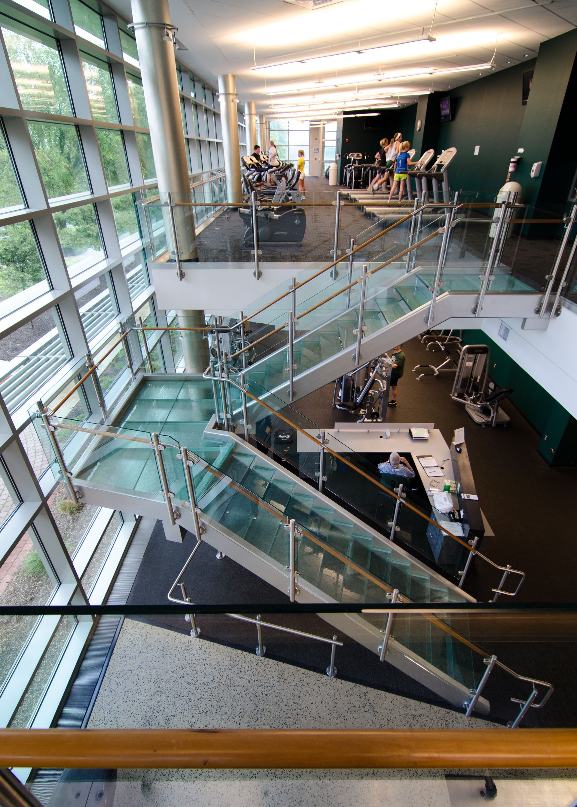 Modern atrium with glass staircases, metal railings, and people gathered on an upper-level walkway