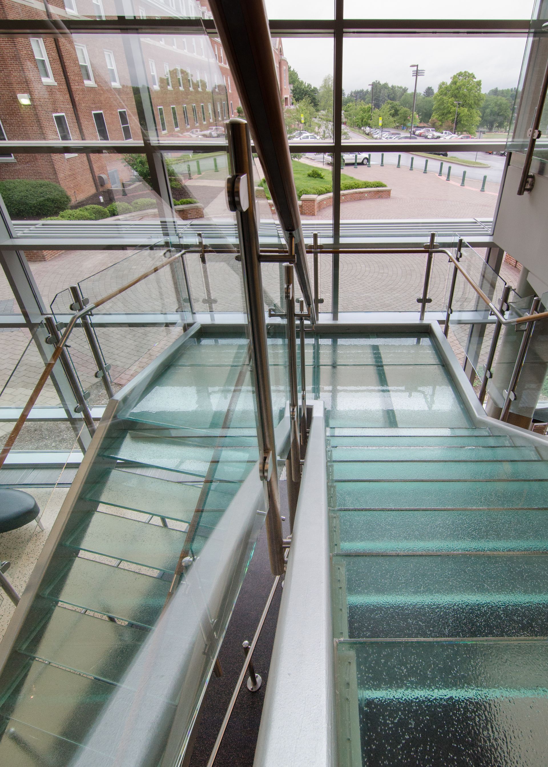 Glass staircase inside a building, overlooking a courtyard and brick campus exterior.