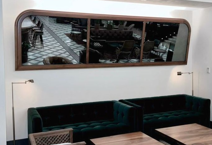 A long, wooden-framed antique mirror over green velvet couches in a lounge area. Two floor lamps provide light.