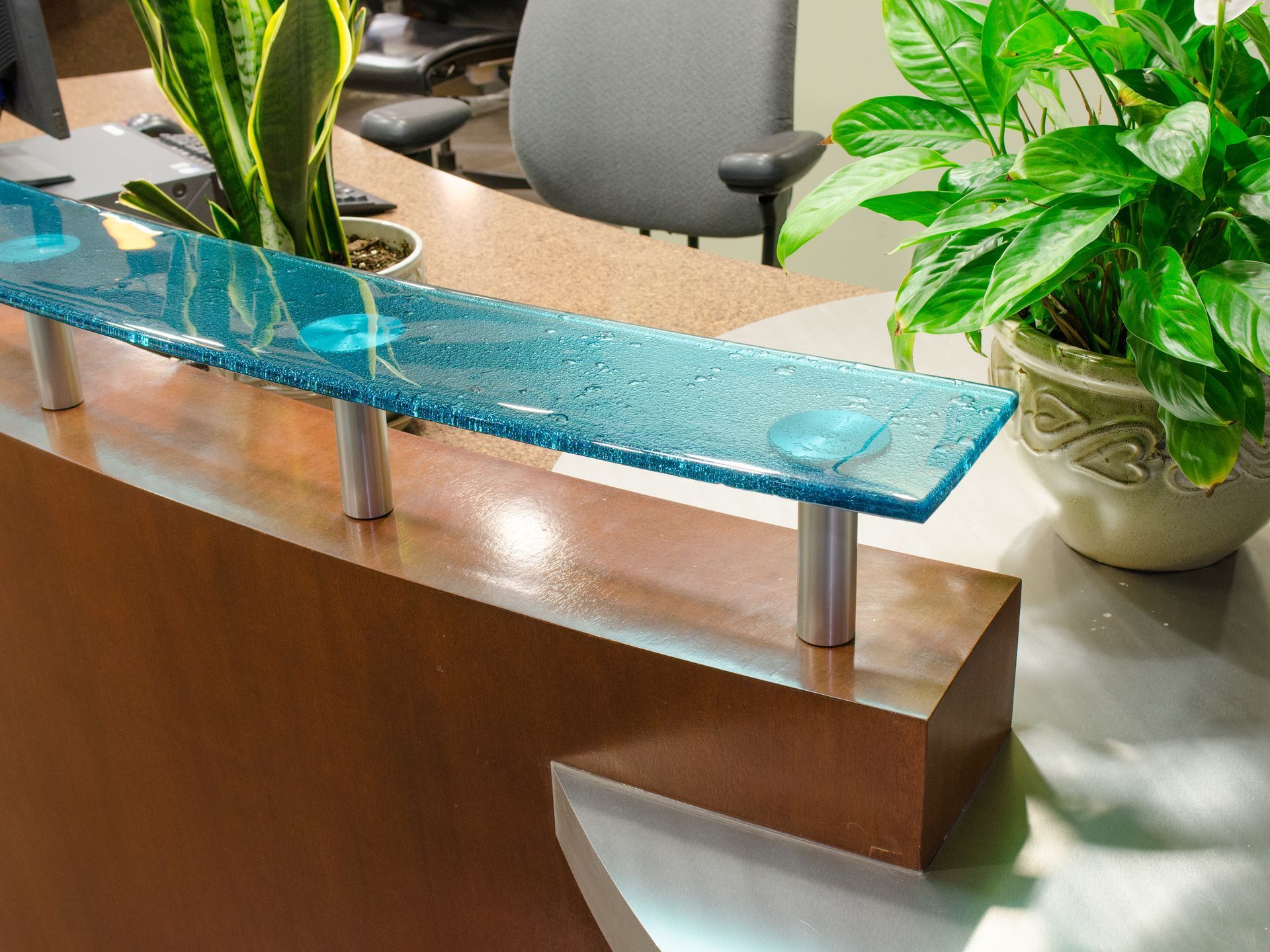 A reception desk with a blue glass top, plants, and a computer in the background.