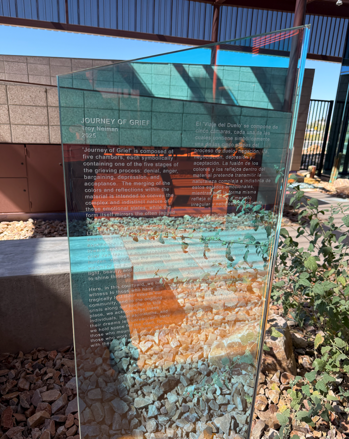 dichroic Glass sign with text, part of a building visible, rocks and plants in foreground.