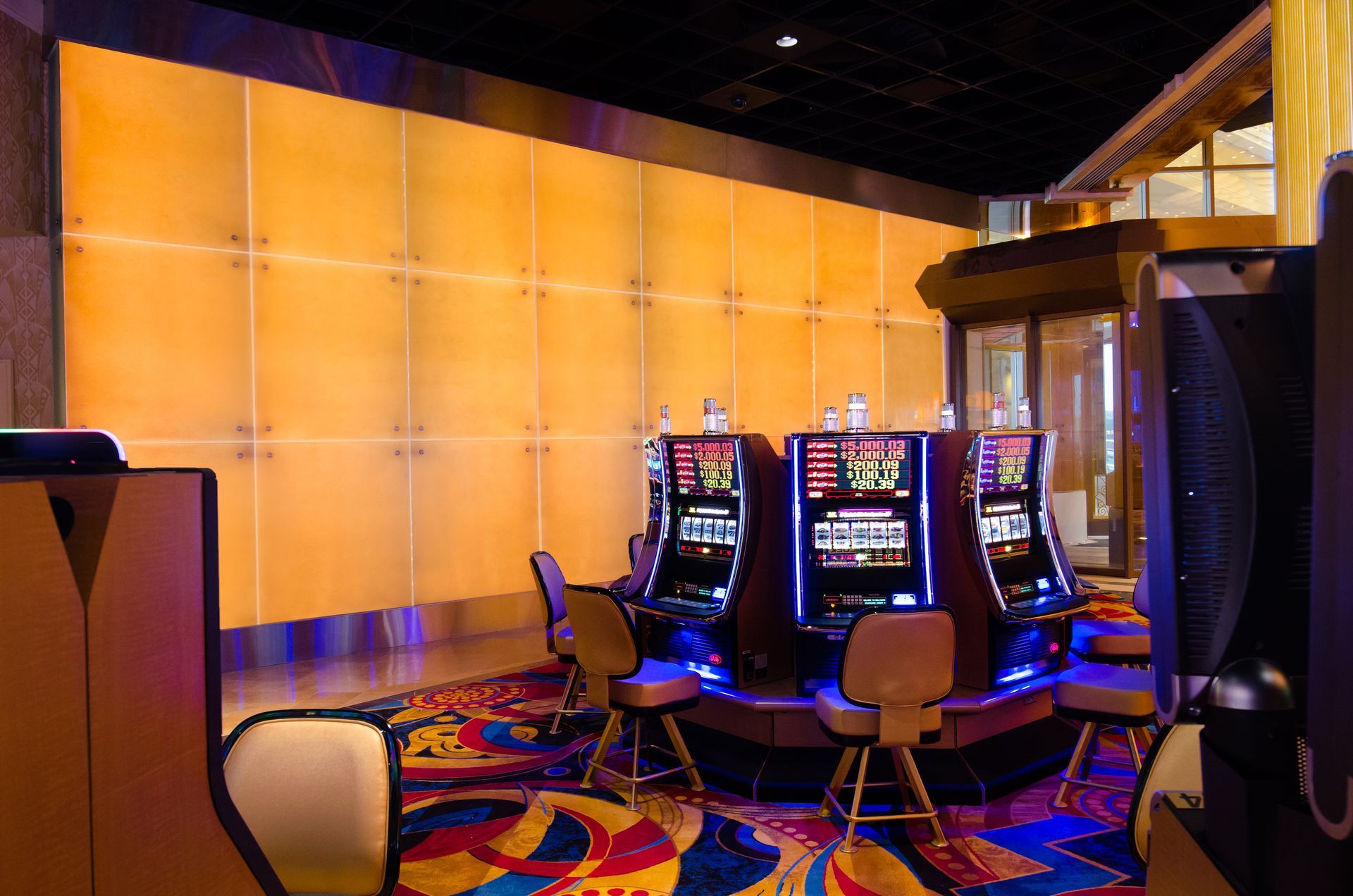 Casino slot machine area with glowing screens, colorful carpet, and empty chairs under warm lighting