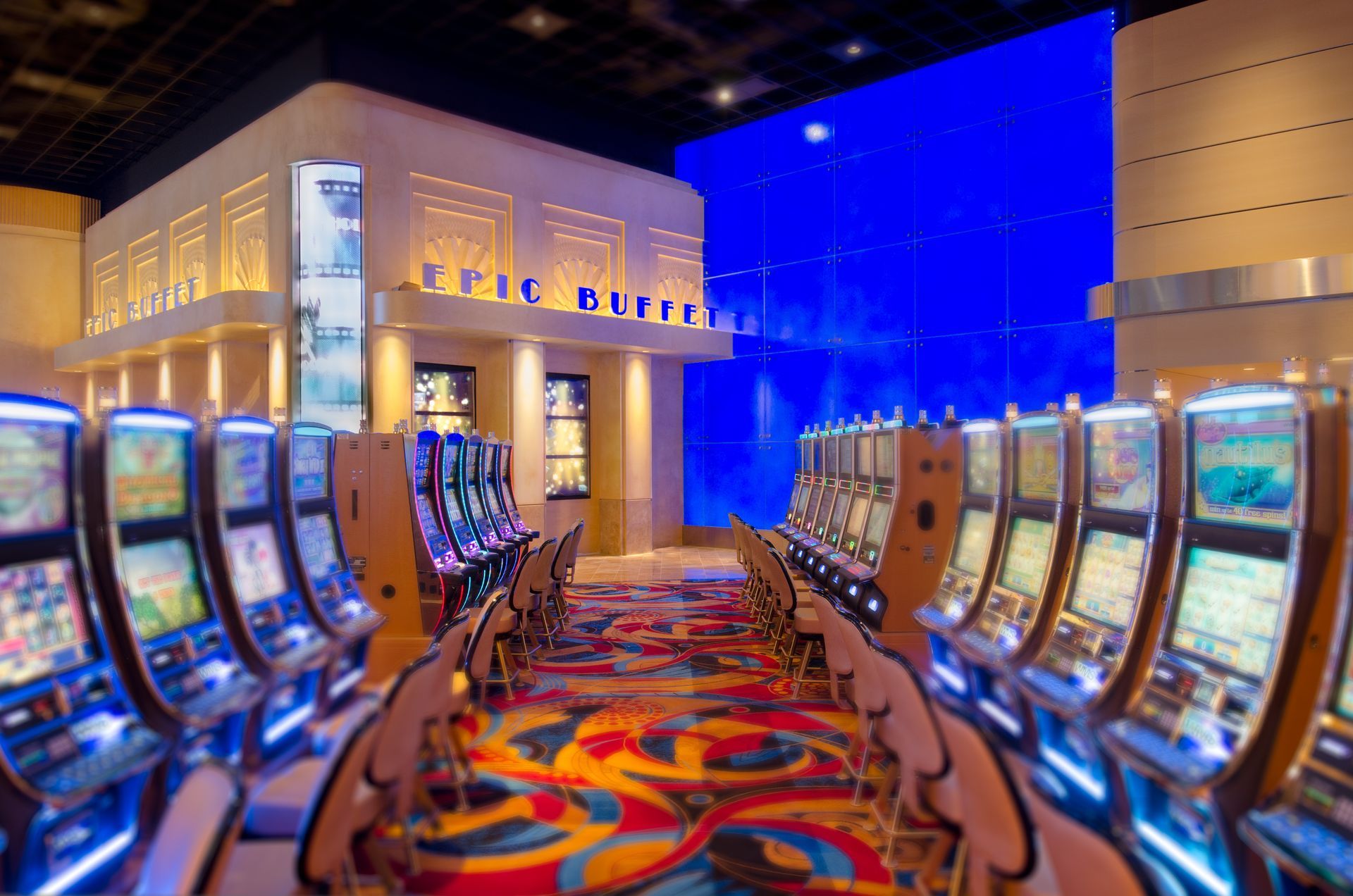 Casino floor with rows of slot machines and a colorful carpet under blue and warm lighting
