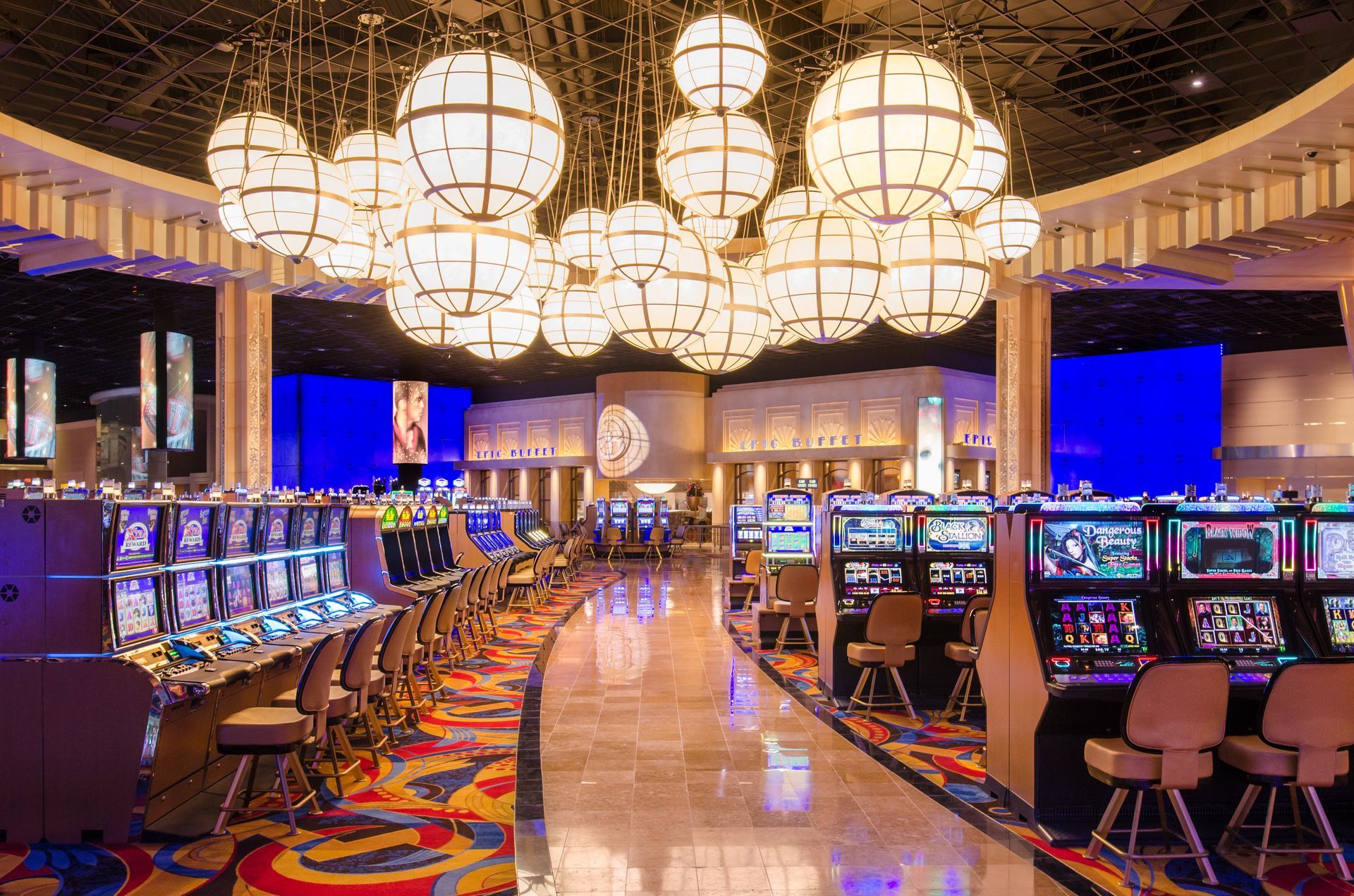 Bright casino floor with glowing slot machines, rows of stools, and large round chandeliers overhead.