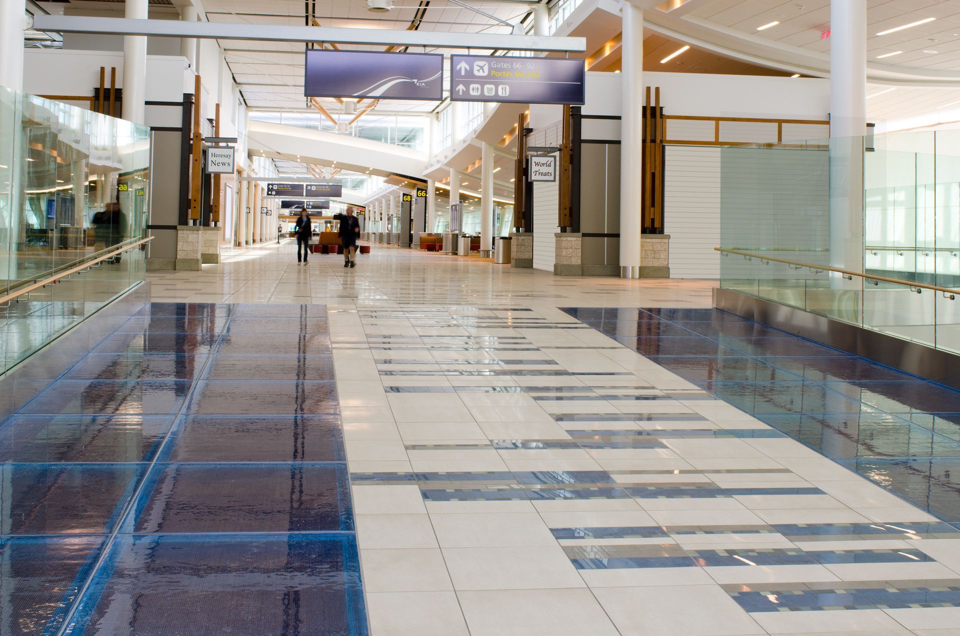 Bright shopping mall corridor with glass railings, blue patterned floor tiles, and a few people in the distance