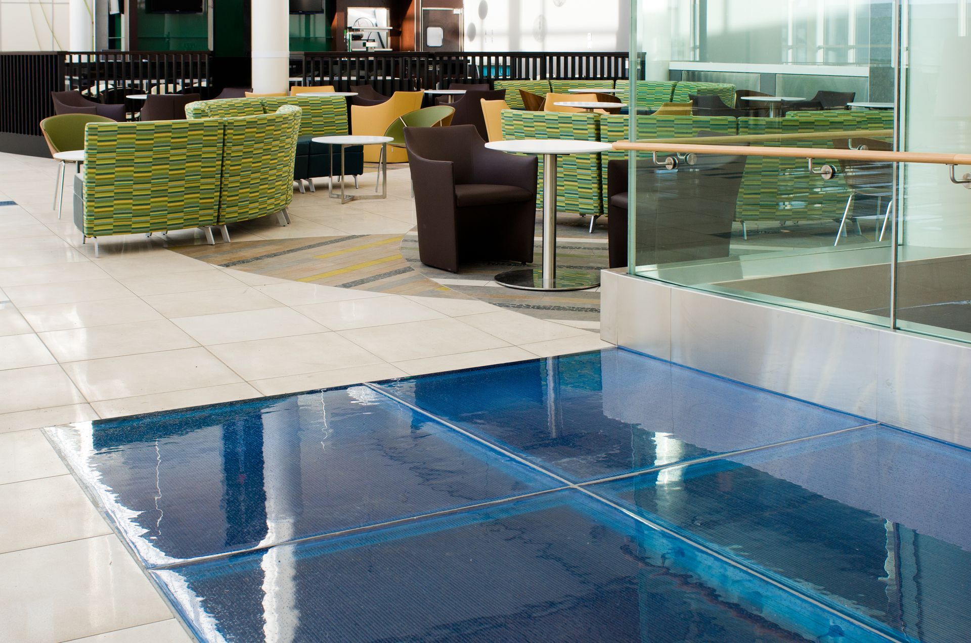 Modern indoor poolside lounge with blue water, green chairs, tables, and glass railings