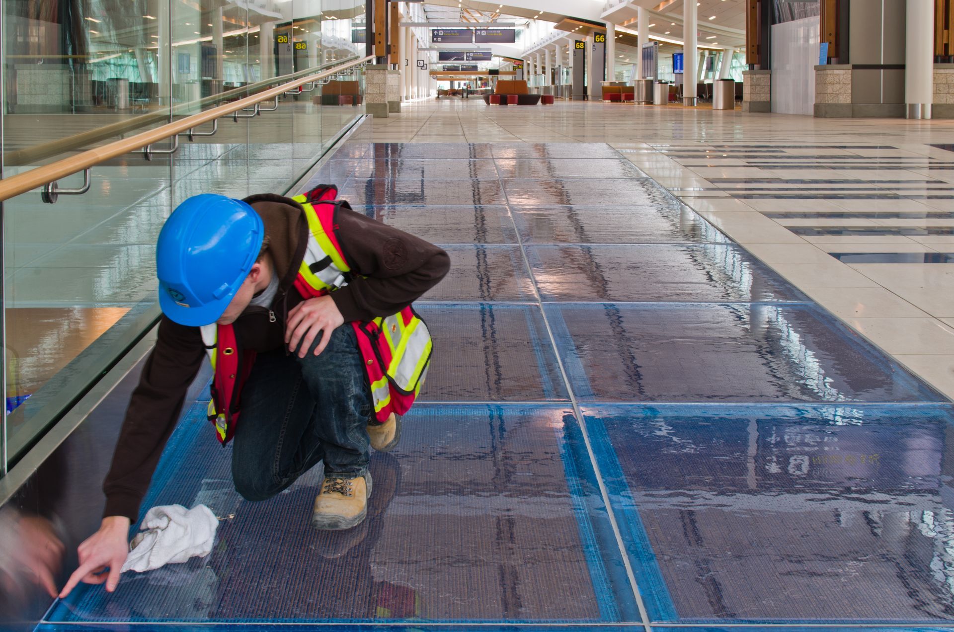Worker in blue hard hat and safety vest inspecting floor tiles in a bright corridor