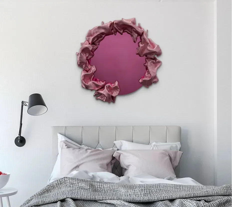 Pink mirror with textured frame above bed with pink pillows and gray blanket.