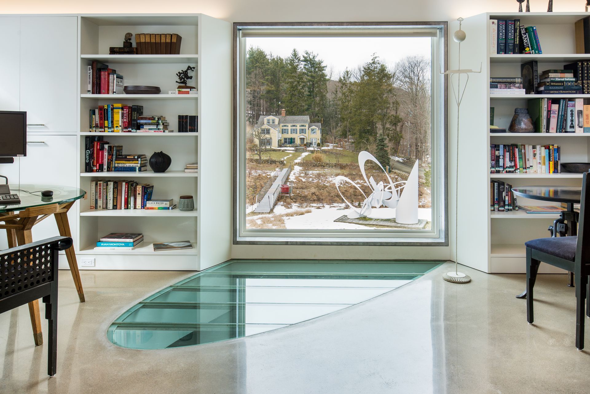 Modern living room with bookshelves, a large window, and a glass floor panel overlooking the outside.