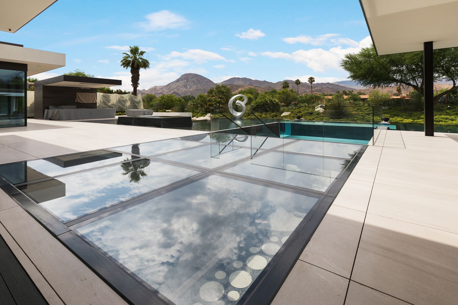 Glass-bottom pool reflects clouds, overlooking a mountainous landscape. Luxurious outdoor setting.