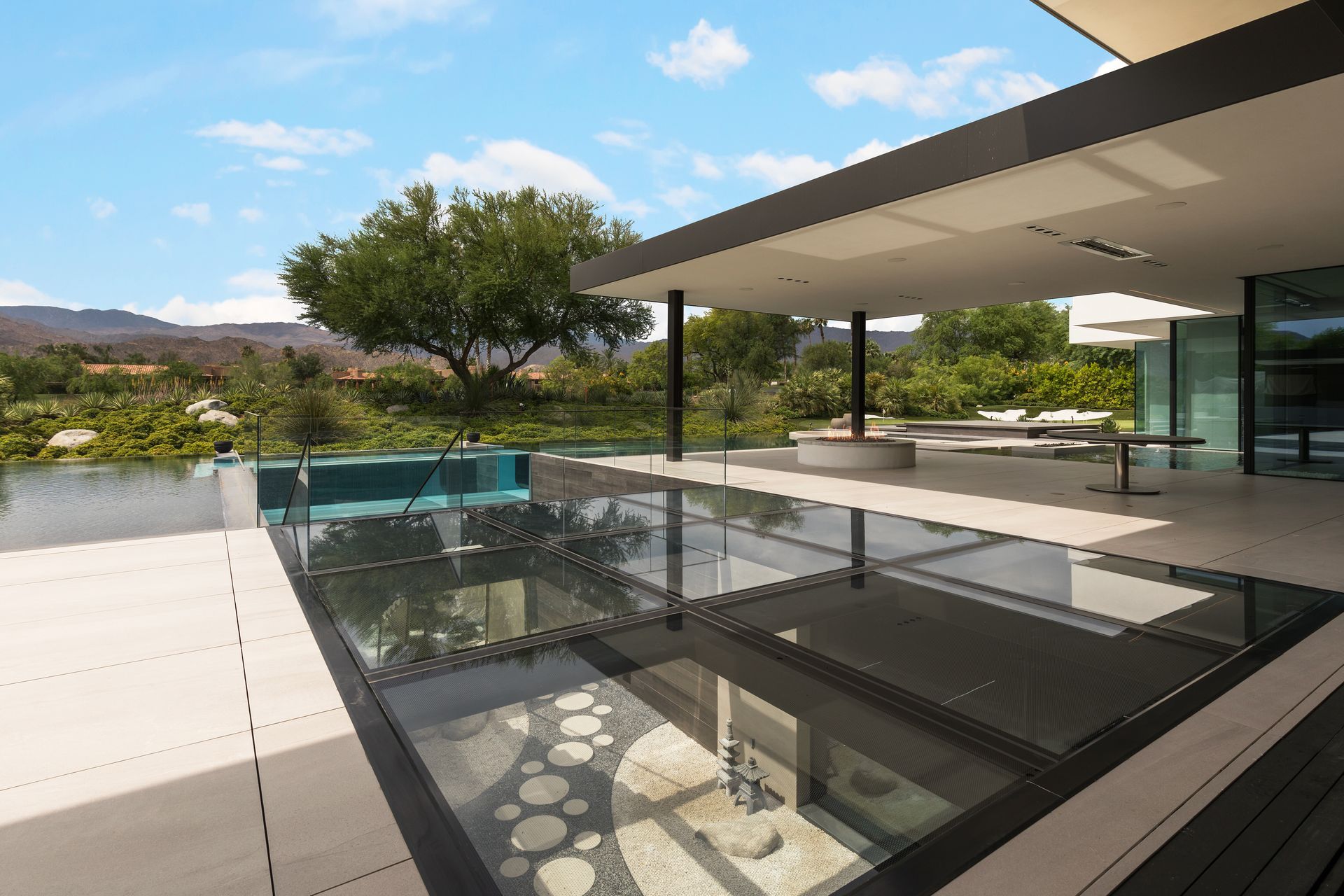 Modern patio with a glass floor looking down to decorative stones; clear pool and greenery in background.