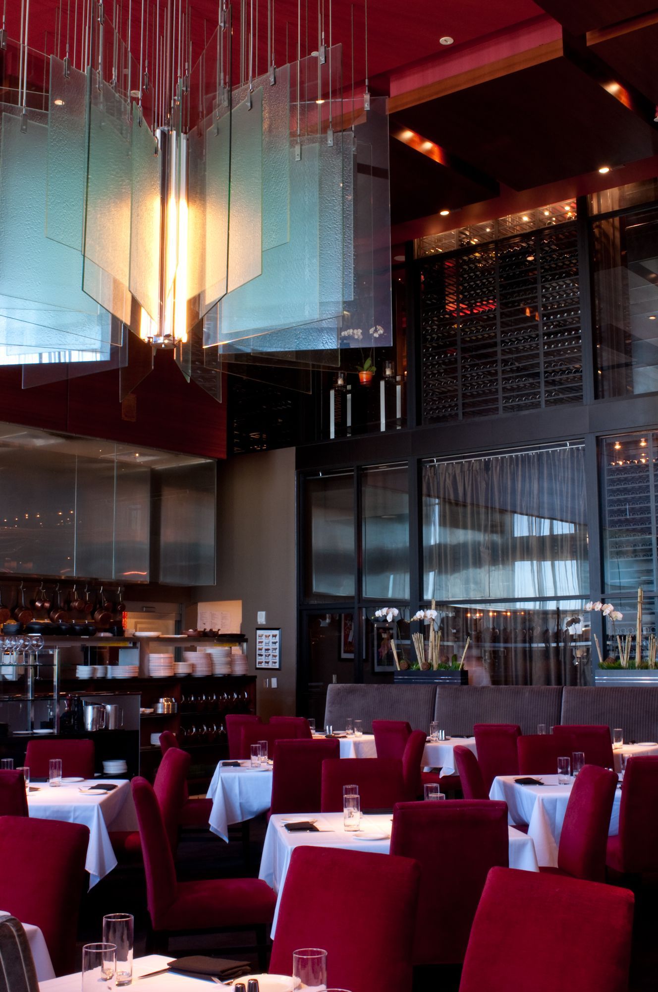 Modern restaurant dining room with red chairs, white tablecloths, and tall glass partitions under warm lighting