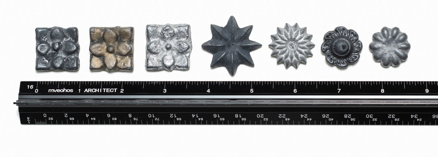 Seven metal decorative rosettes of various shapes and shades arranged in a row above a standard black measuring ruler.
