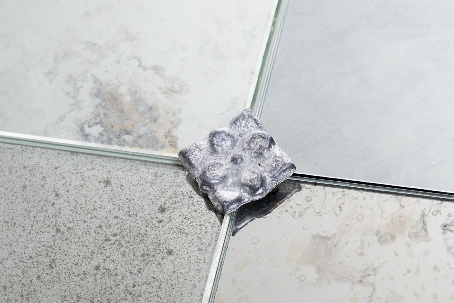 A small, textured, silver-colored decorative accent piece sits at the intersection of four mirrored glass tiles.