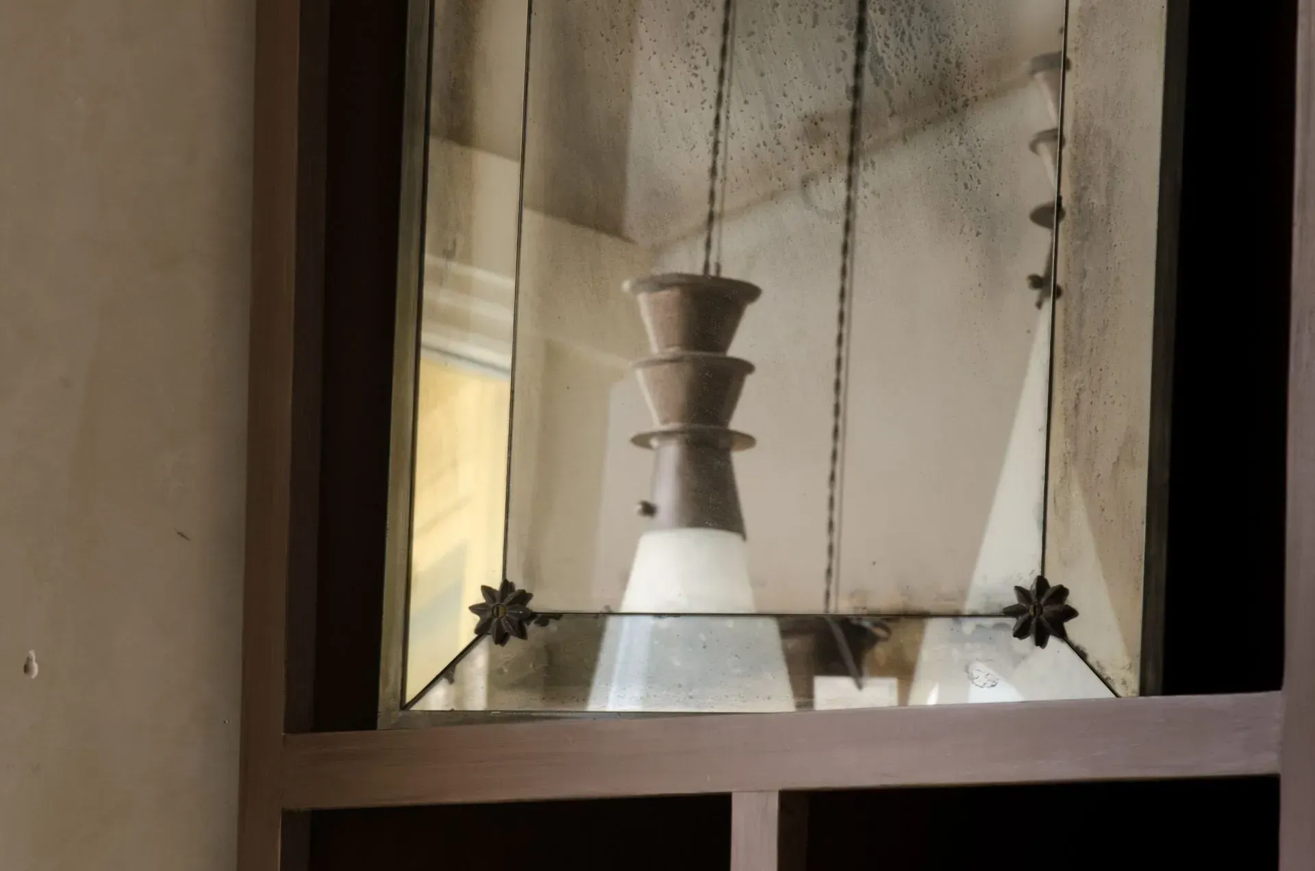 A tiered, grey pendant light with a frosted glass shade, seen reflected in a framed mirror with decorative corner details.