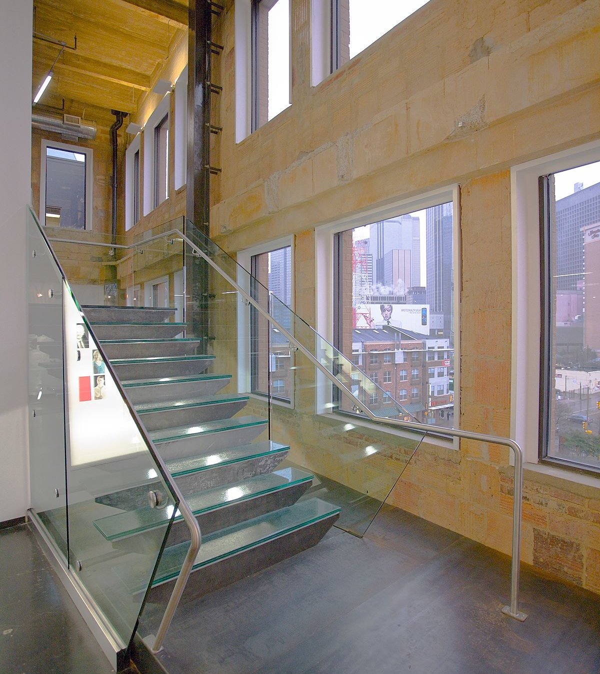 Modern staircase with glass treads and railings in a bright building with tall windows showing a city view.
