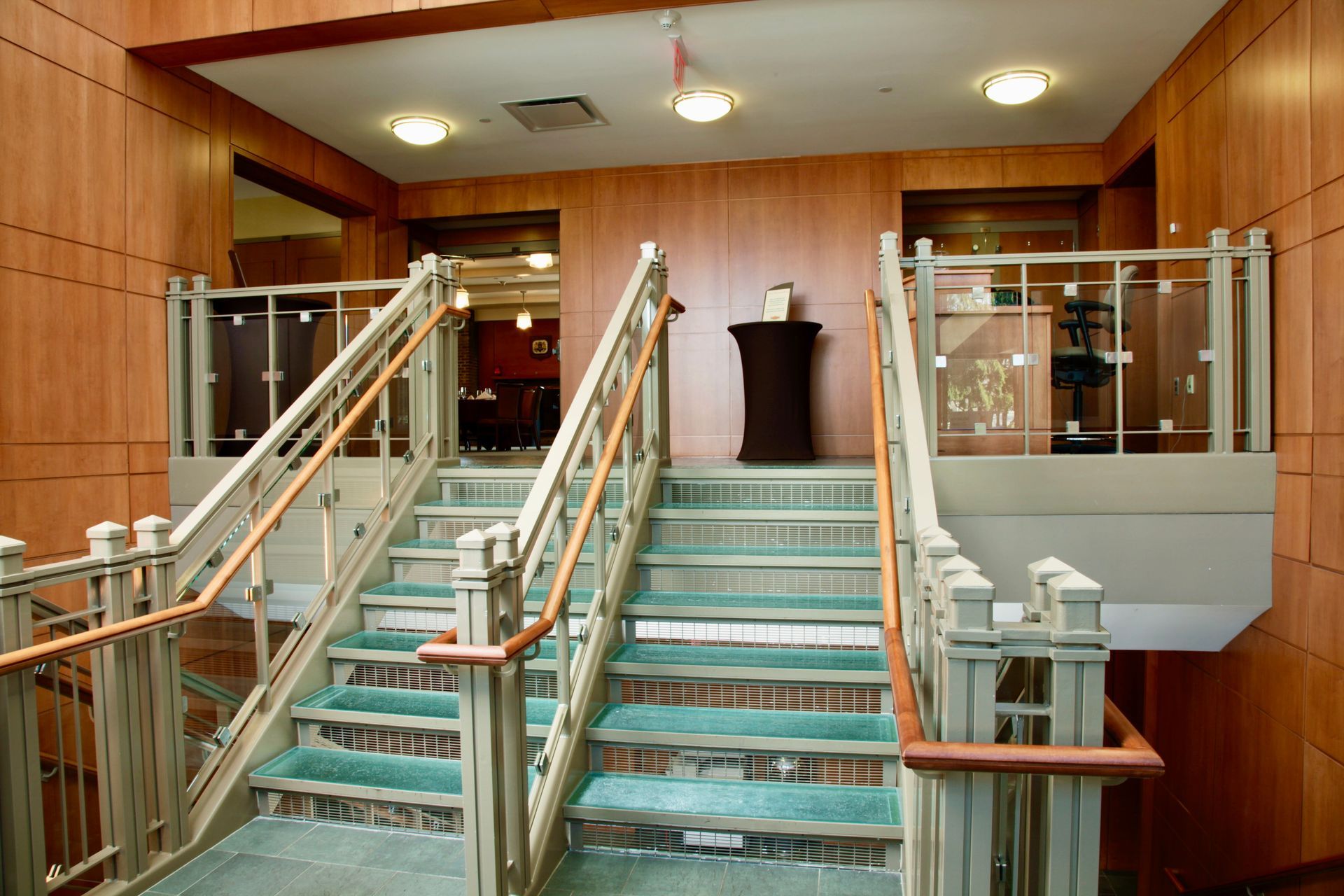 A set of stairs with light-colored railings leads up to a landing in an indoor hallway with wood-paneled walls.