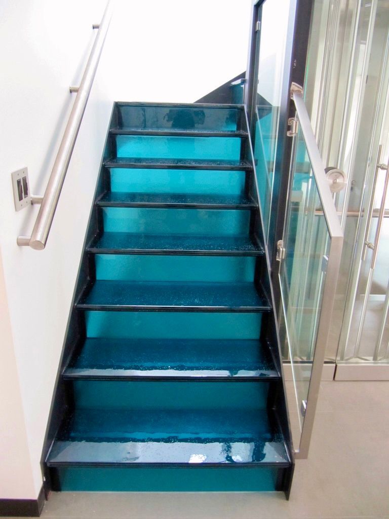 Staircase with turquoise glass treads and black borders, with a metal handrail on one side and a glass panel on the other.