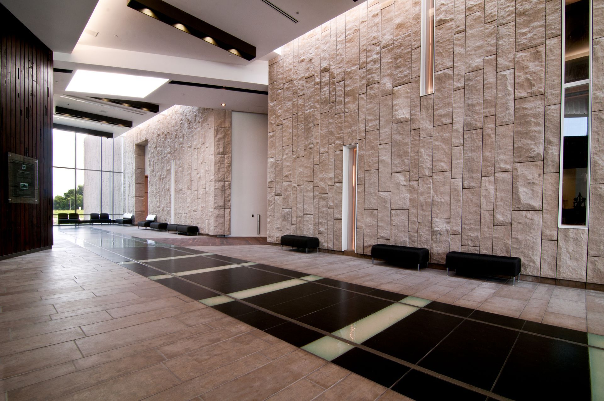 Modern lobby corridor with tall stone walls, black benches, and tiled flooring under skylights