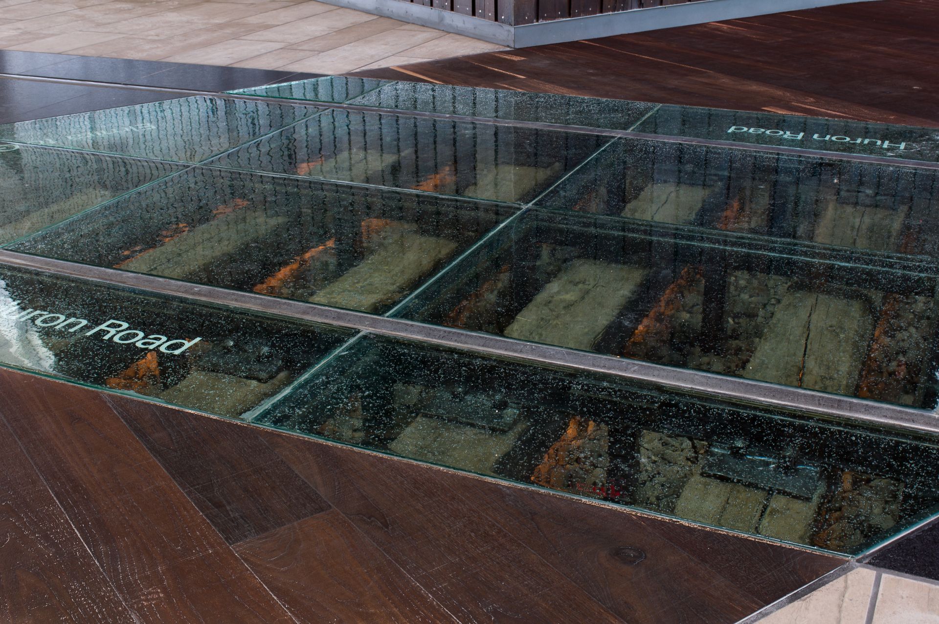 Glass-floor view of a lower level with display cases and wooden flooring, seen through panes with reflections