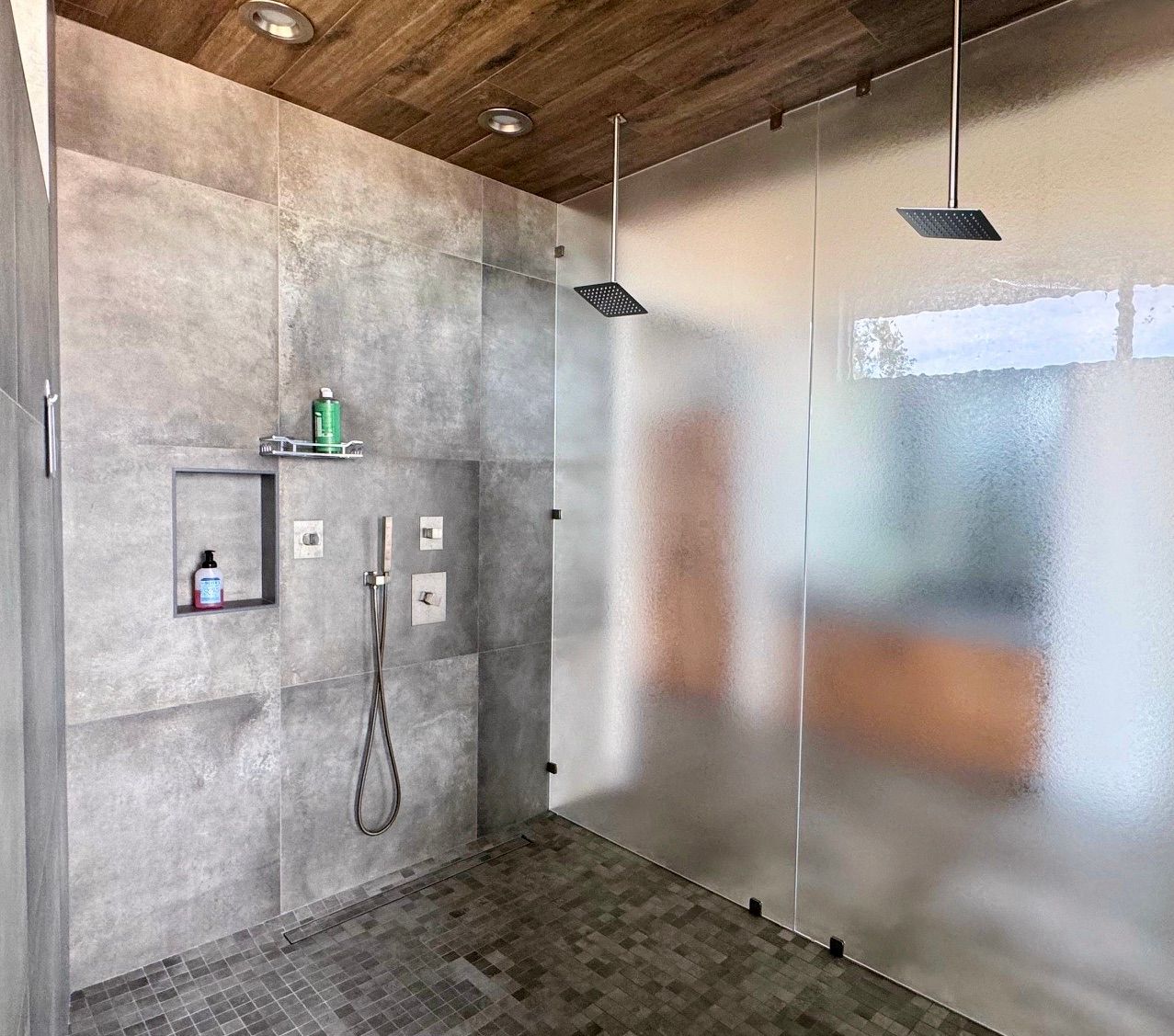 Large modern shower with gray tile walls, textured glass door, and two shower heads. Wooden ceiling.