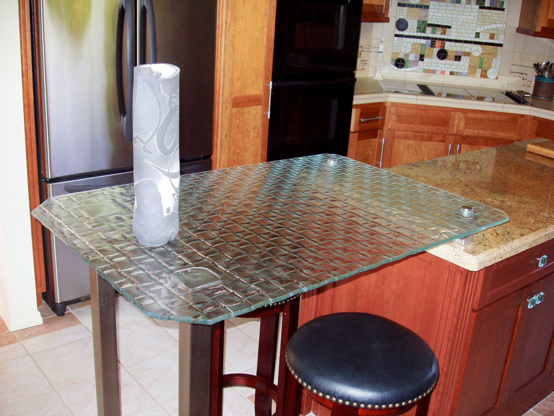 Cast Glass-top kitchen island with a vase on it, and a stool. Dark cabinetry and granite countertop are visible.