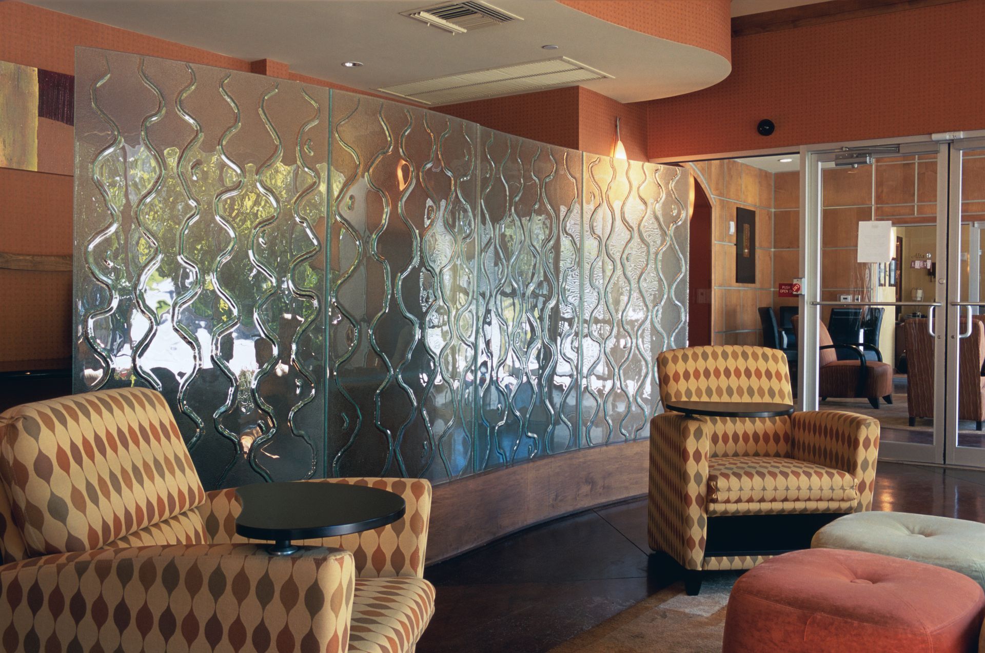 Lounge area with textured glass wall, two patterned armchairs, and colorful ottoman seating.