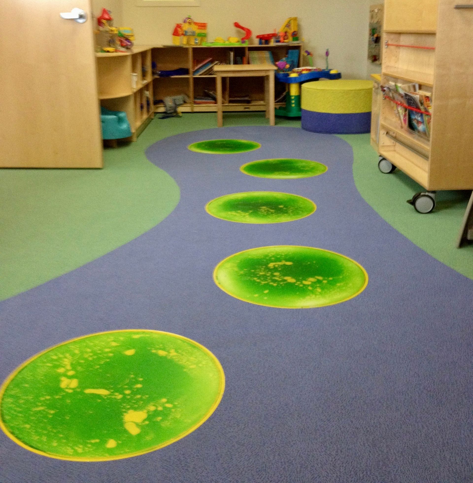 Playroom floor featuring a blue path with green lily pad-style circles leading toward shelves, toys, and small furniture.