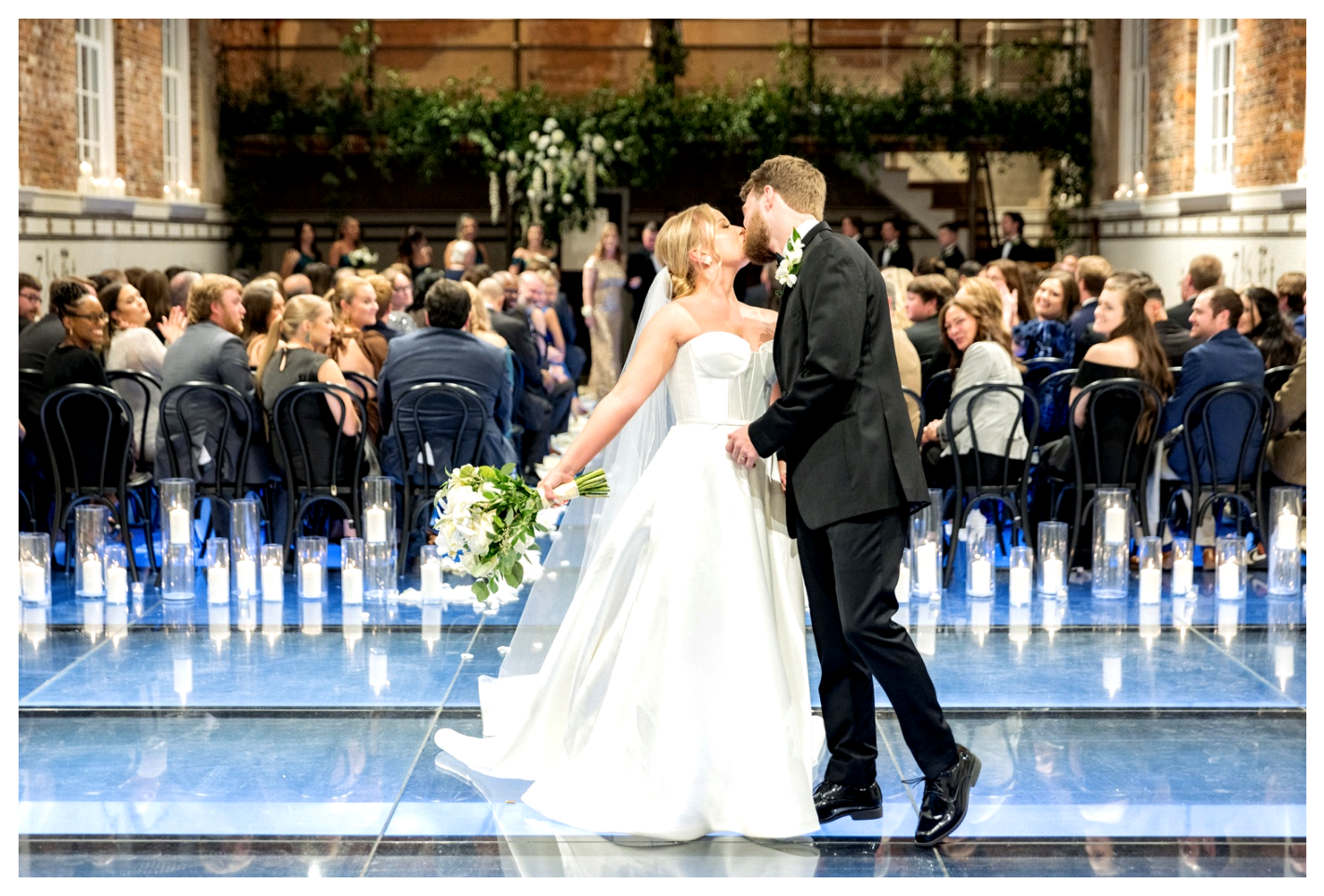 Bride and groom kissing in a grand church with guests seated behind them, reflections on the blue floor