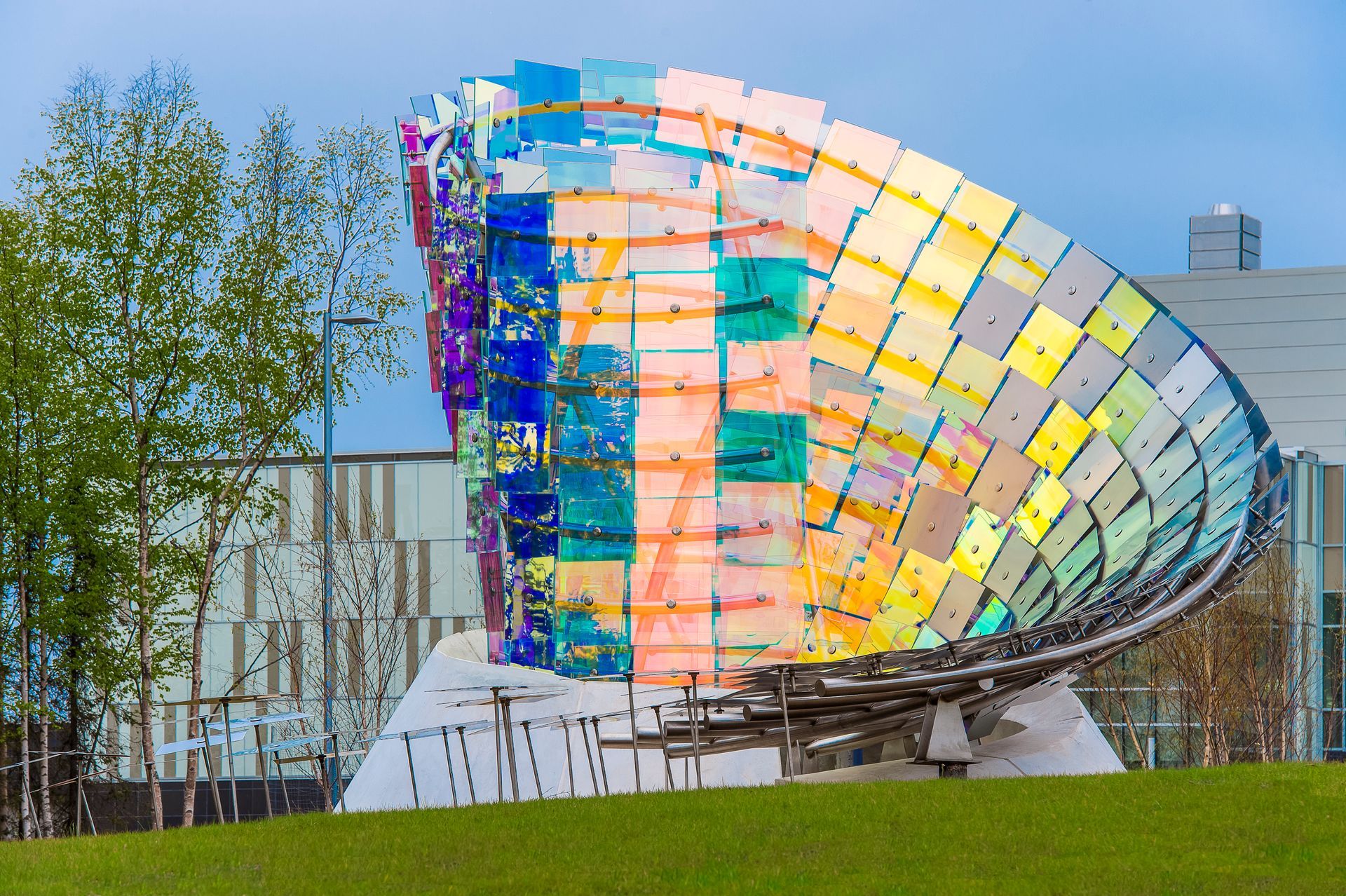 Colorful glass sculpture resembling a giant shell in a grassy outdoor park with trees and buildings behind it