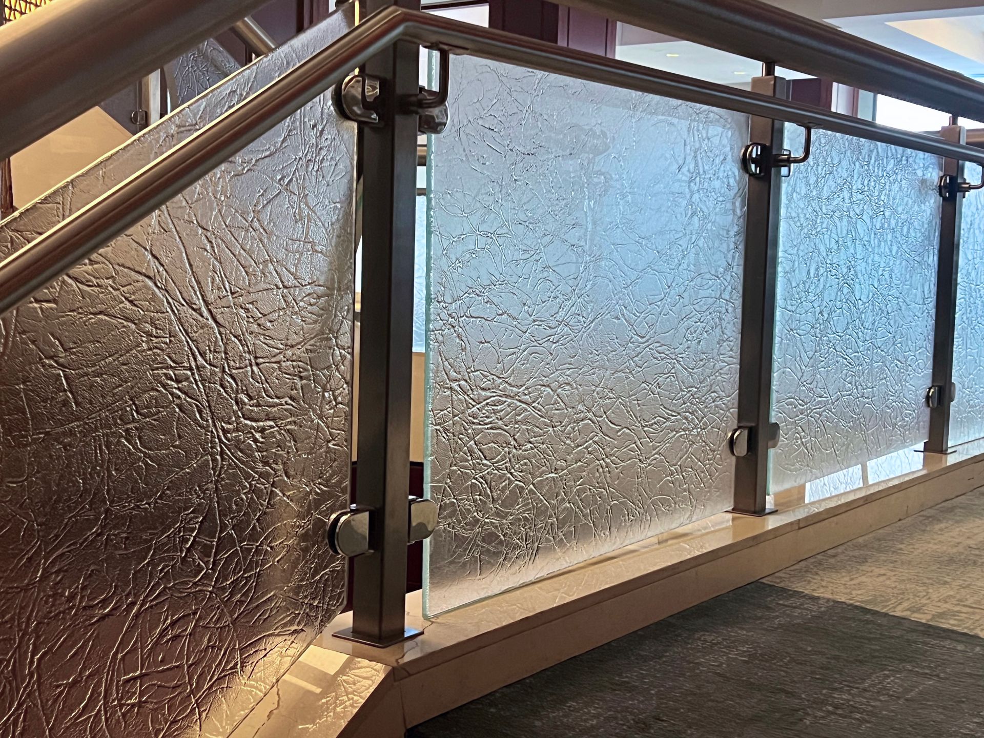 Glass railing with textured surface, stainless steel supports, and wooden base along a staircase.