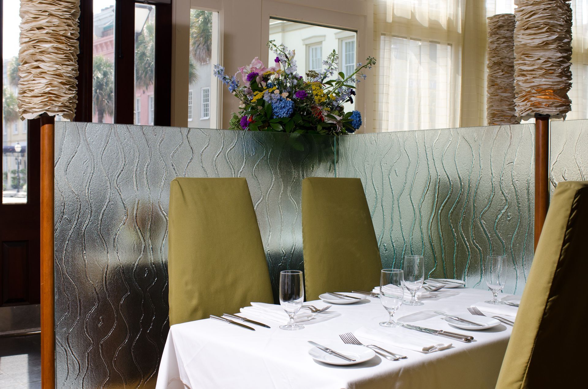 Elegant restaurant table by frosted glass partition, with green chairs and a floral centerpiece.