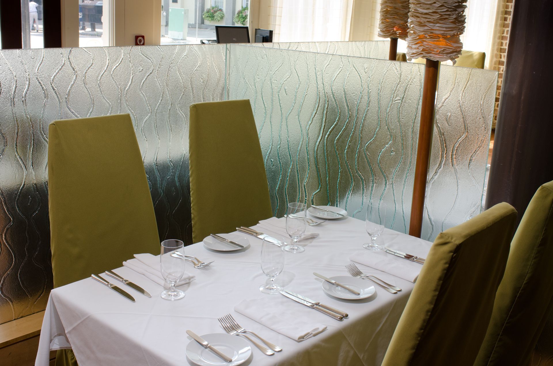 Restaurant table set with white tablecloth, glassware, and green chairs beside a frosted window.