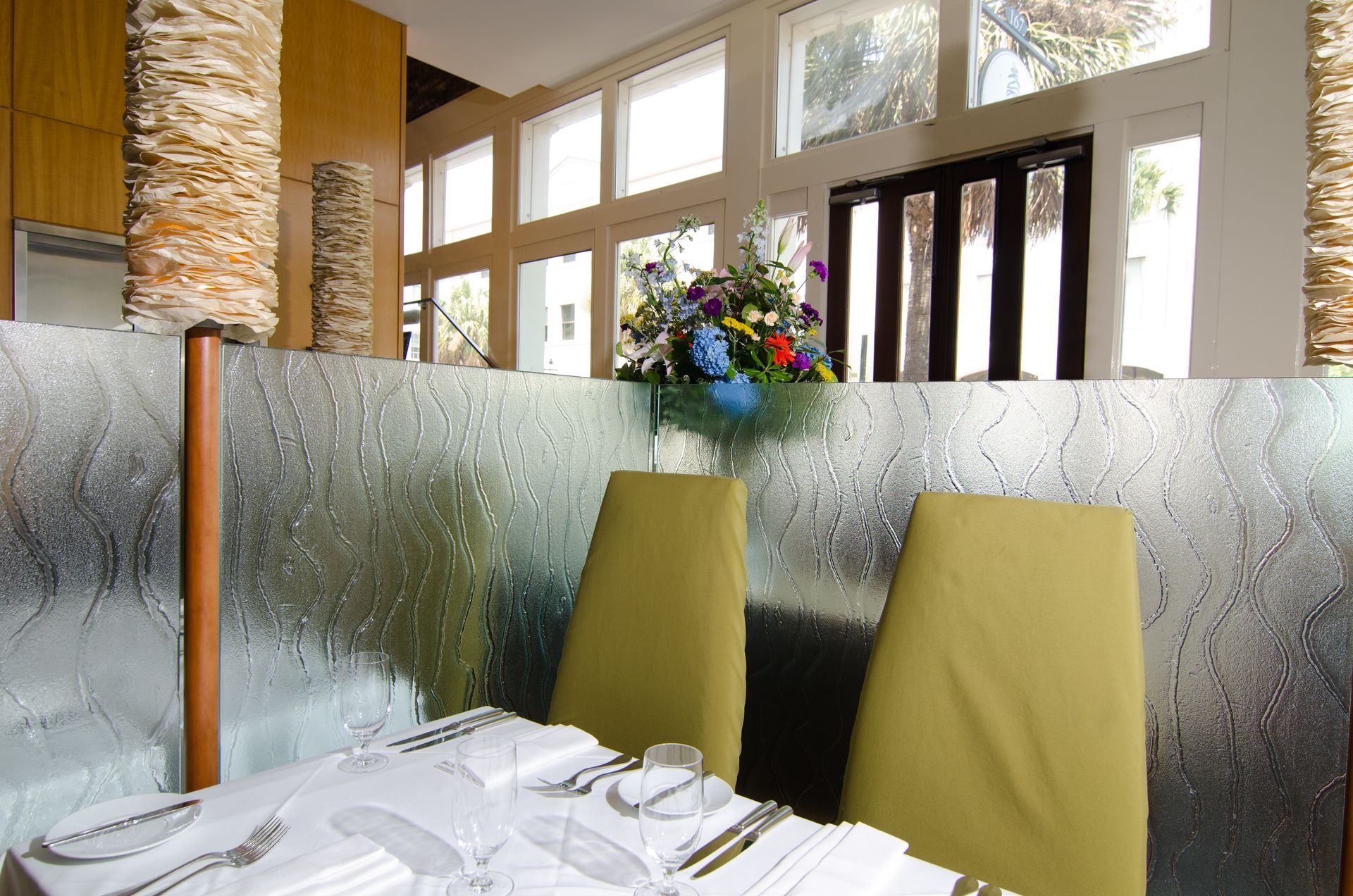 Restaurant table with white napkins, two yellow chairs, frosted glass partition, and a flower arrangement by tall windows