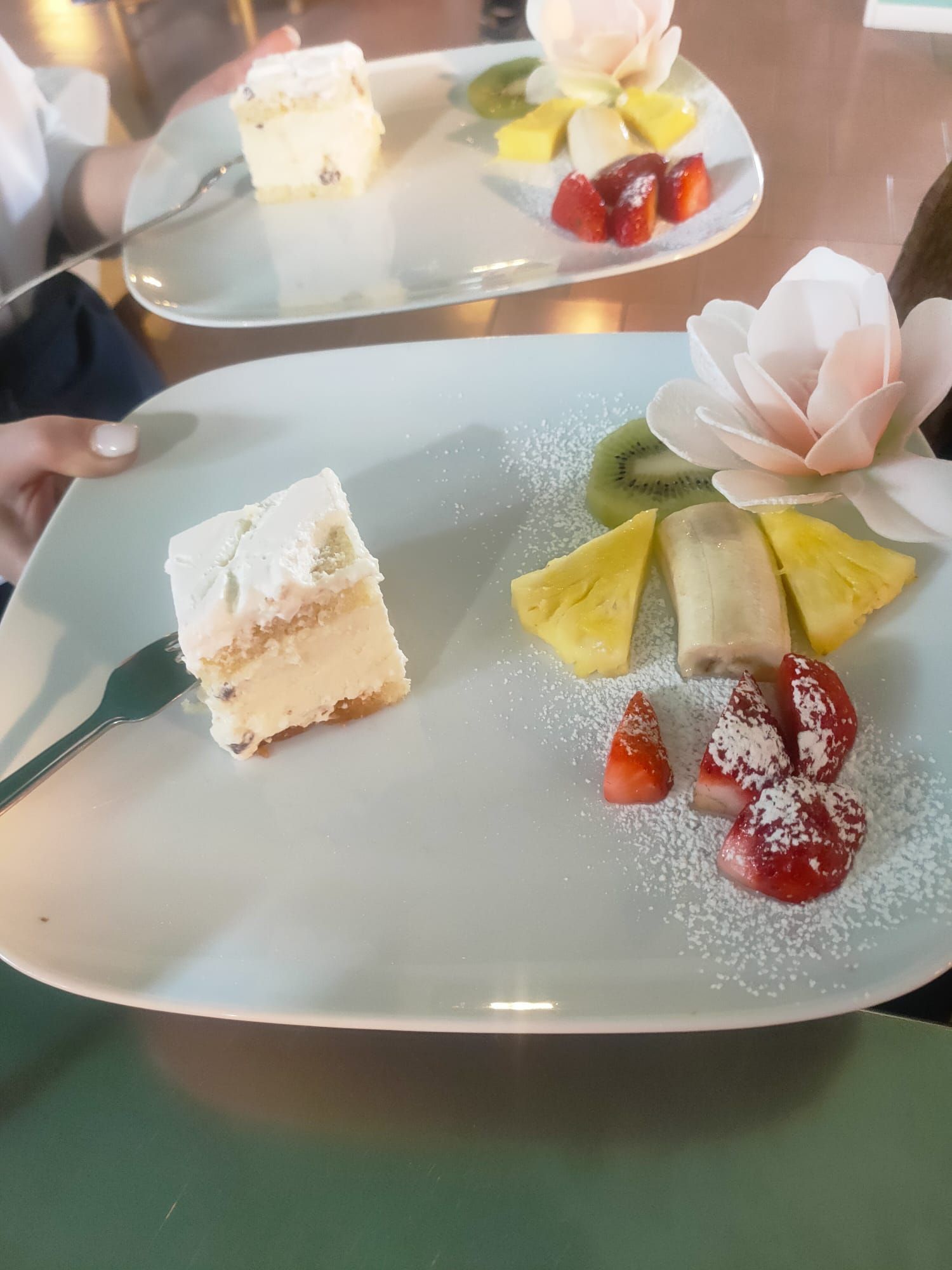 Due fette quadrate di torta a strati servite su piatti bianchi con una guarnizione di ananas, banana, kiwi, frutti di bosco e un fiore.