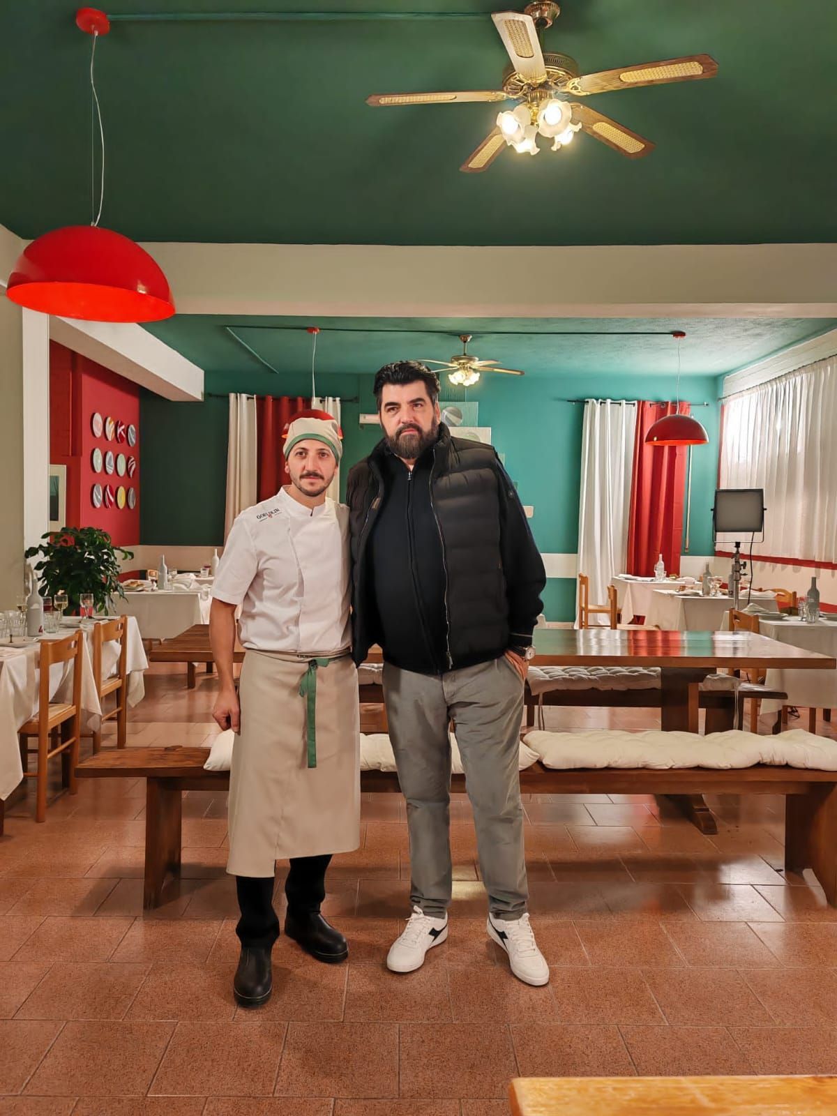 Due persone posano per una foto in un ristorante rustico con pareti verdi, un ventilatore a soffitto e mobili in legno.