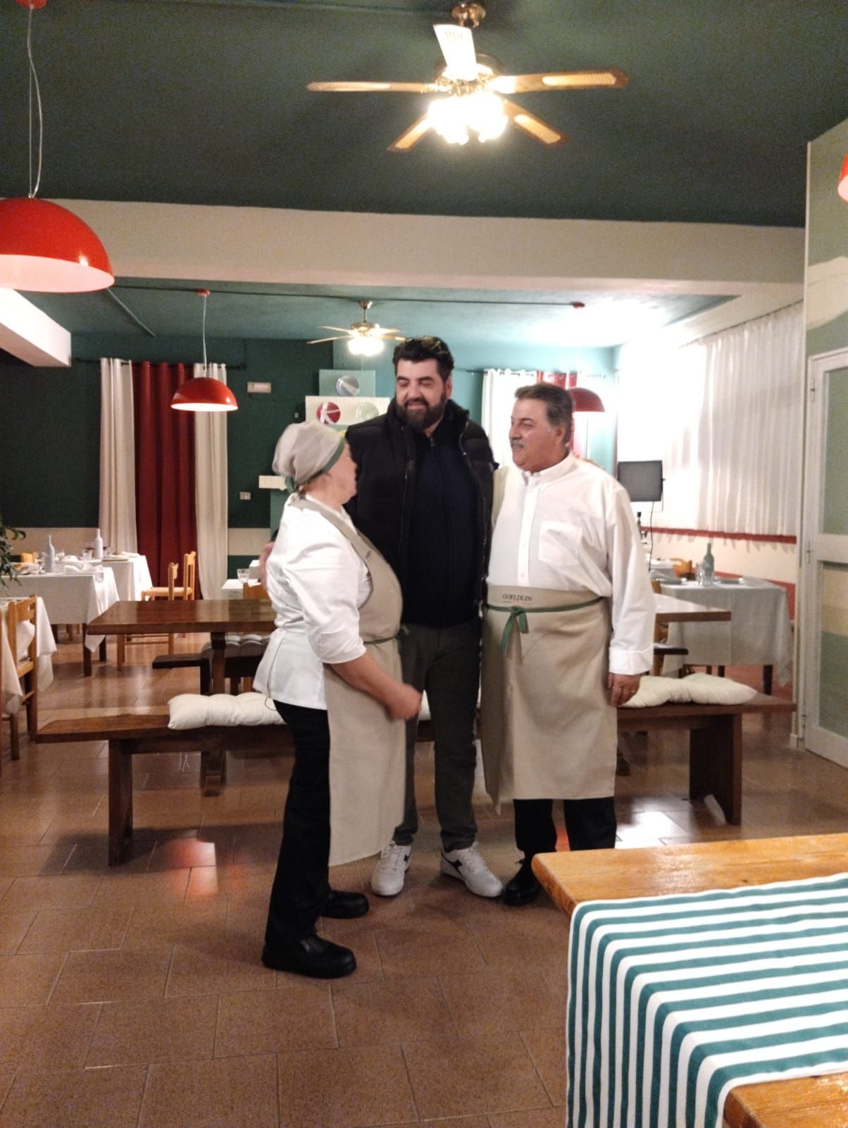In un ristorante, due persone con il grembiule sono in piedi: una è rivolta verso un uomo con una giacca nera, mentre l'altra osserva la scena.