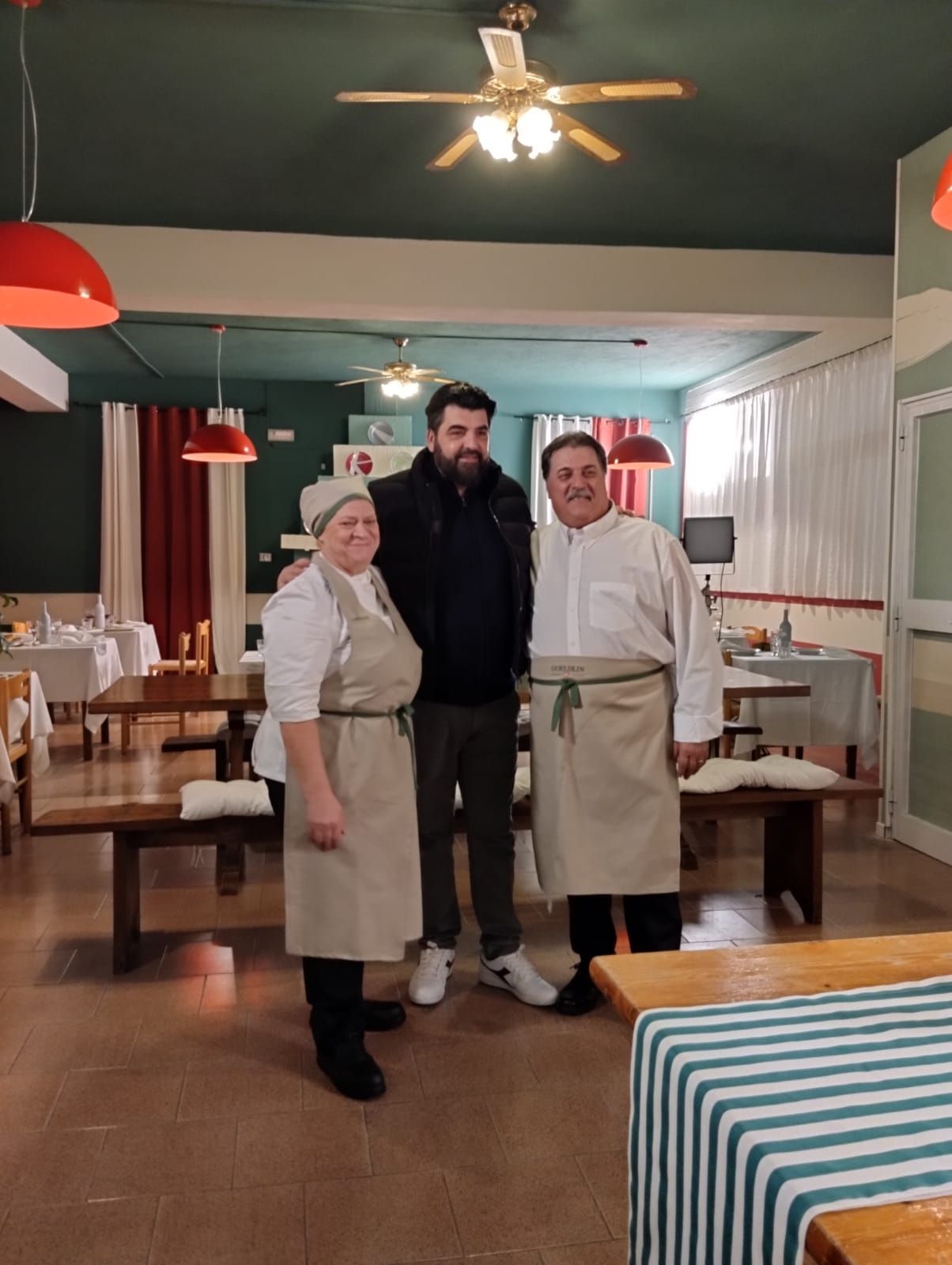 In una sala da pranzo, uno chef e un membro del personale, in uniforme e grembiule bianchi, sono in piedi accanto a un uomo in abiti casual.