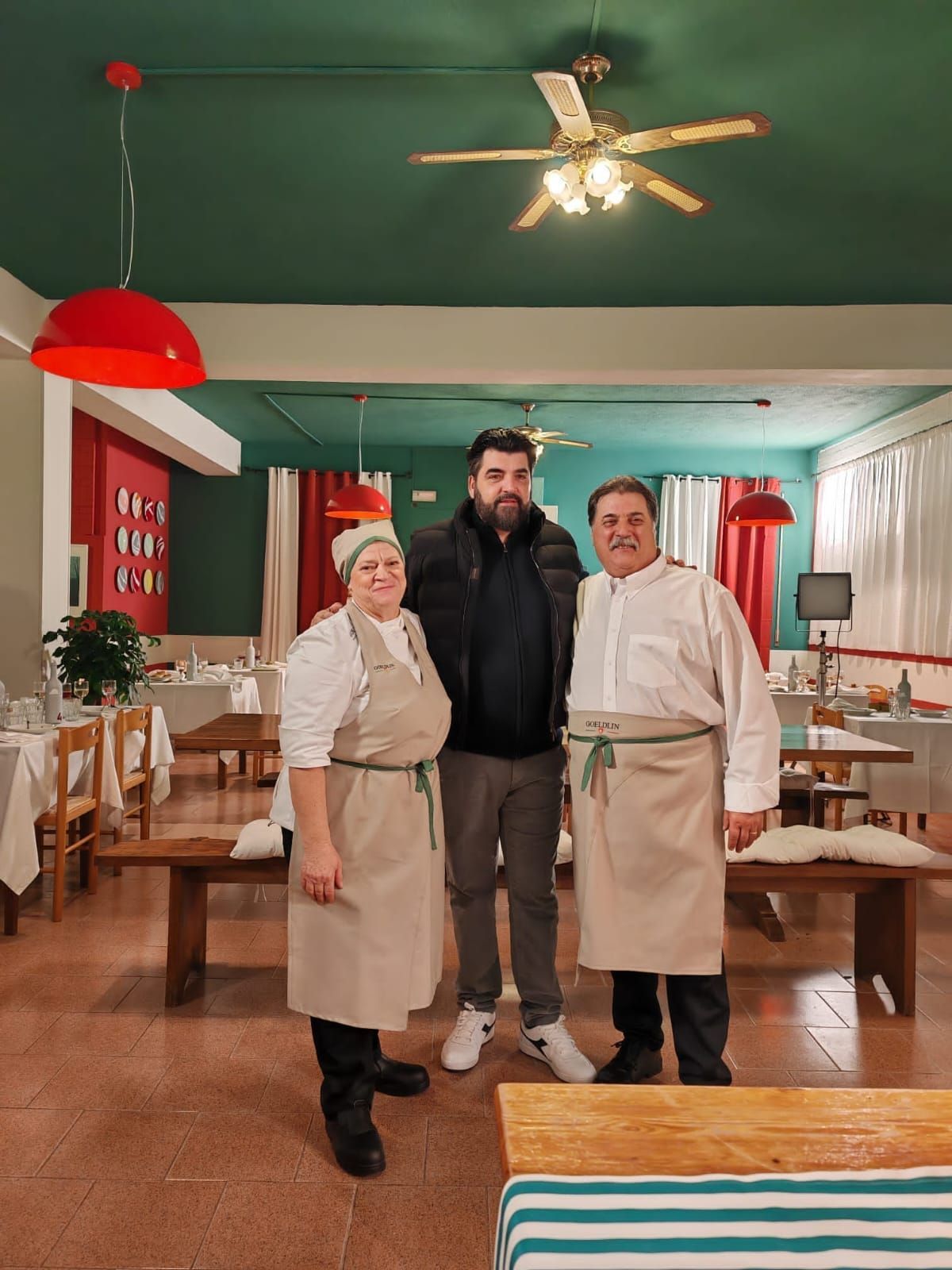 Un uomo sorridente con un giubbotto imbottito posa con una donna e un uomo in uniforme da ristorante all'interno di un accogliente locale dalle pareti verdi.