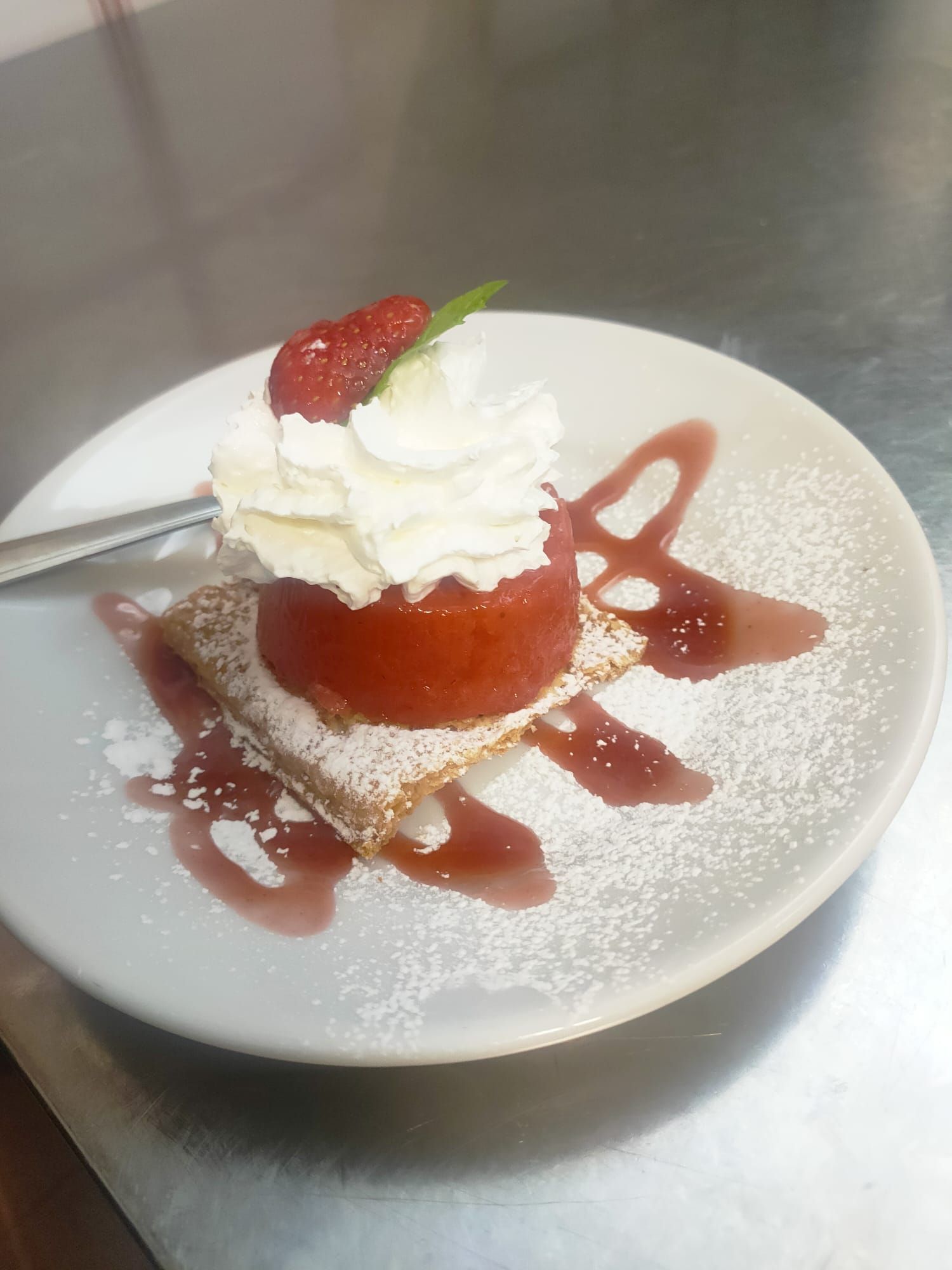 Un dessert al piatto con un frutto rosso sciroppato di forma circolare su un quadrato di pasta sfoglia, guarnito con panna montata e sciroppo.