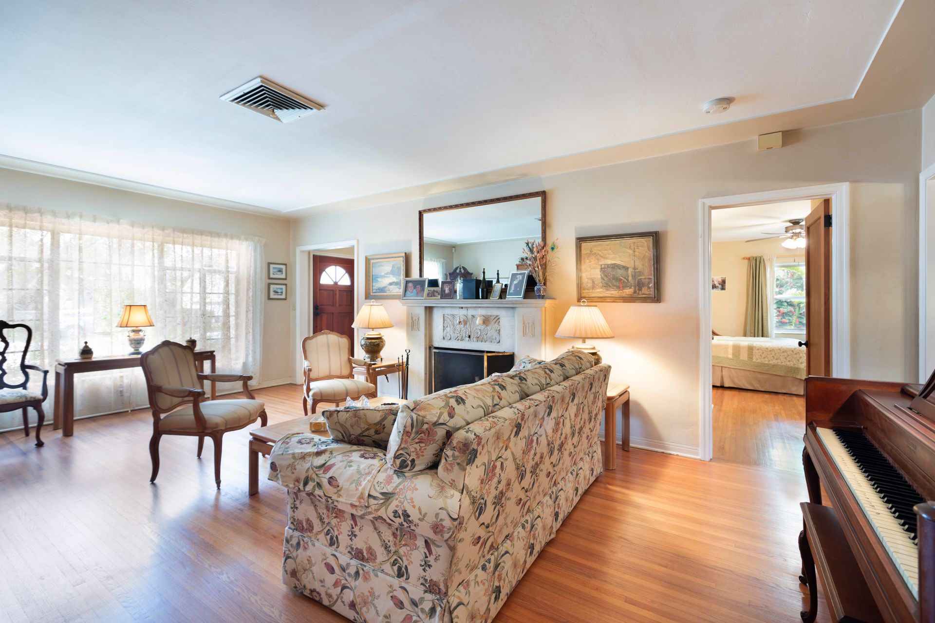 A bright, traditional living room with a floral sofa, armchair, piano, and views into an adjacent room and doorway.