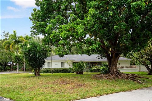 A white, single-story ranch house sits in a grassy yard, partially obscured by a large, leafy tree with exposed roots.