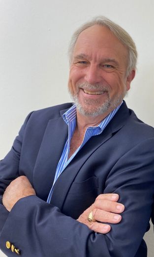 A person with a beard and gray hair smiles, wearing a navy blazer and a blue-and-white striped shirt, arms crossed.