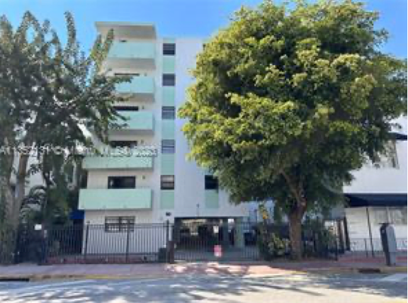 A five-story residential building with light green balconies, bordered by trees and a metal fence on a sunny day.