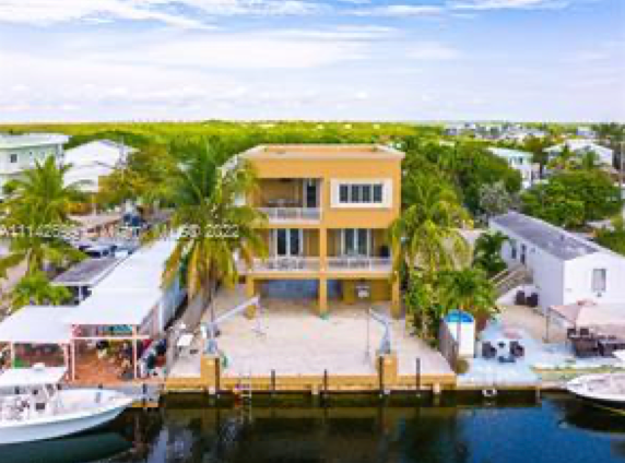 A tan three-story waterfront home with a balcony and dock, situated on a canal surrounded by tropical trees.