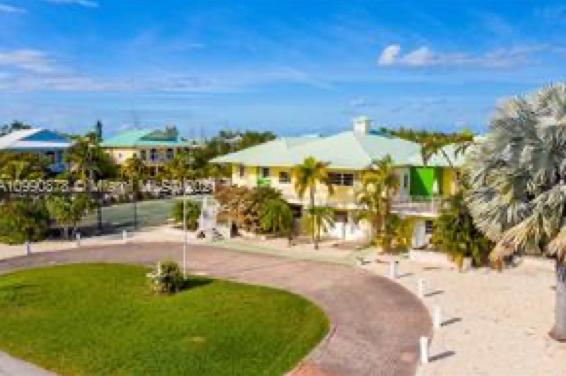 A yellow house with a light-green roof, surrounded by palm trees, a green lawn, and a curved driveway on a sunny day.