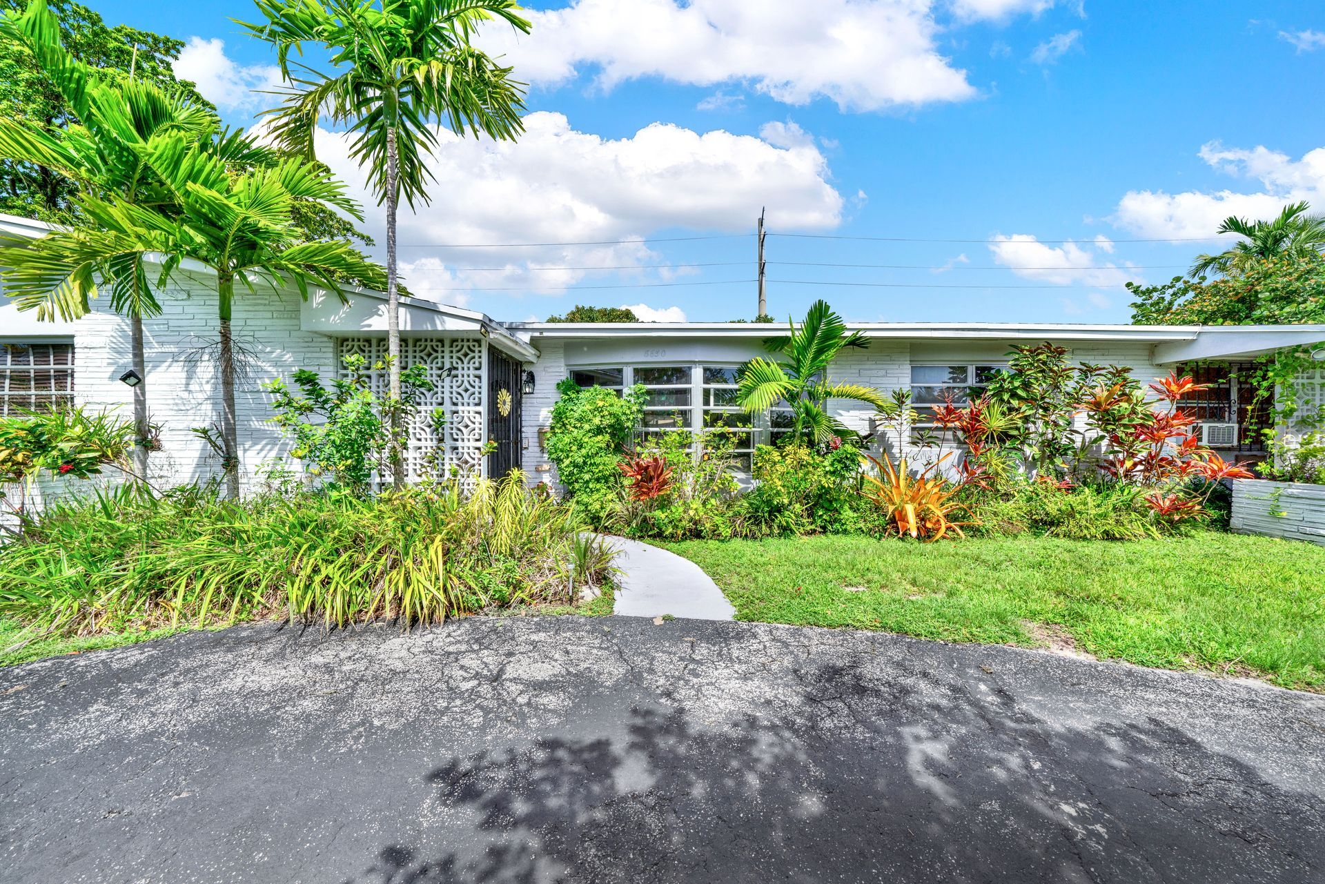 A one-story white house with a large front yard, palm trees, and lush landscaping under a bright blue, cloudy sky.