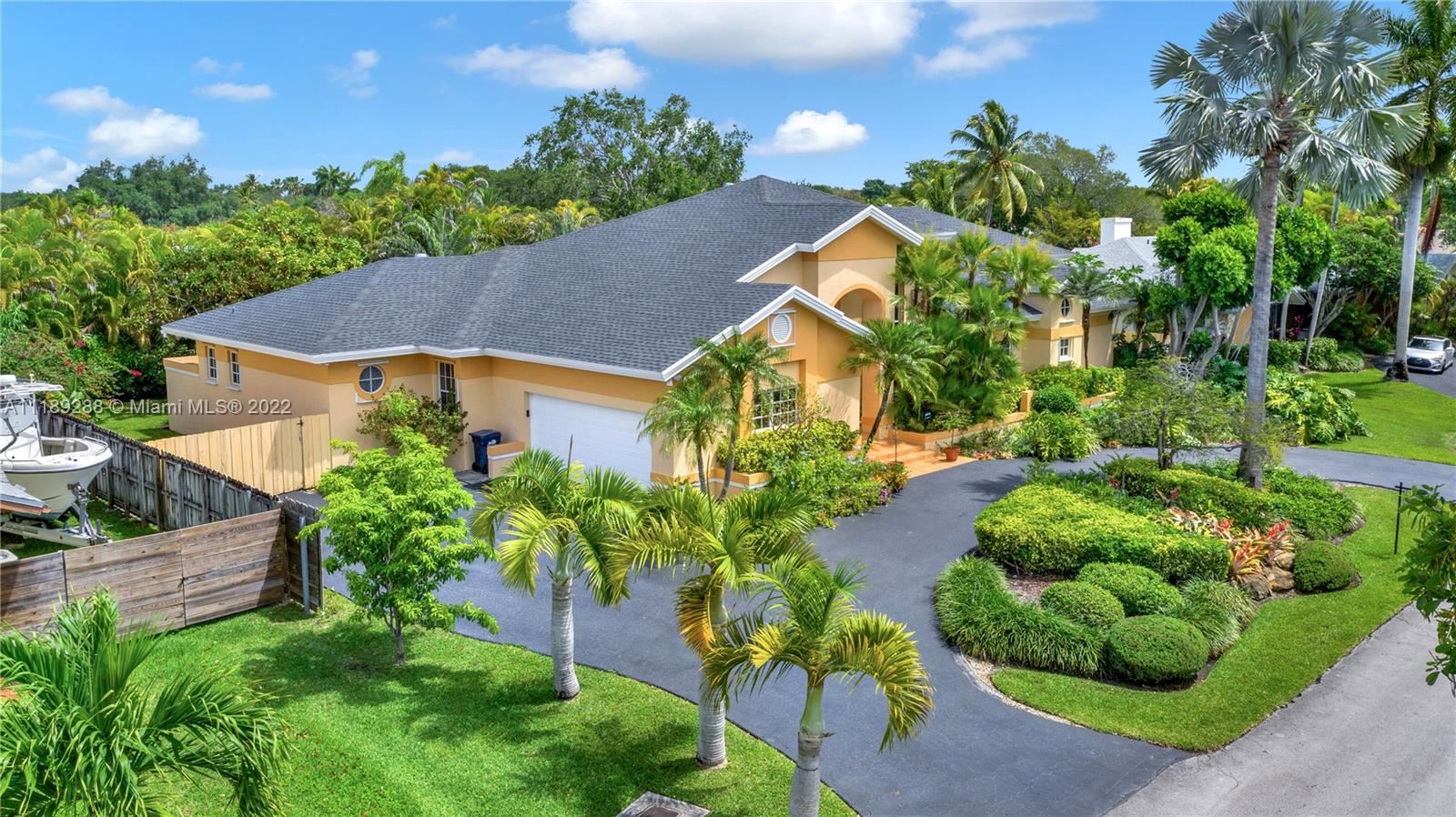 An aerial view of a tan, single-story suburban house with a dark gray shingled roof, palm trees, and a curved driveway.