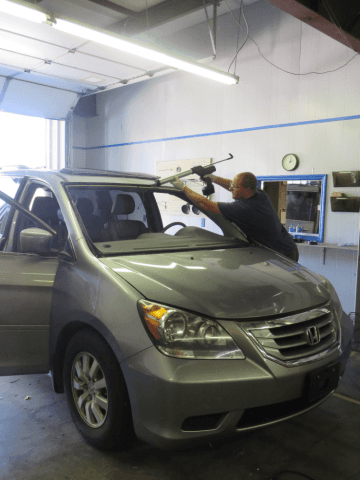 Mechanic removing windshield from a silver minivan inside a garage.